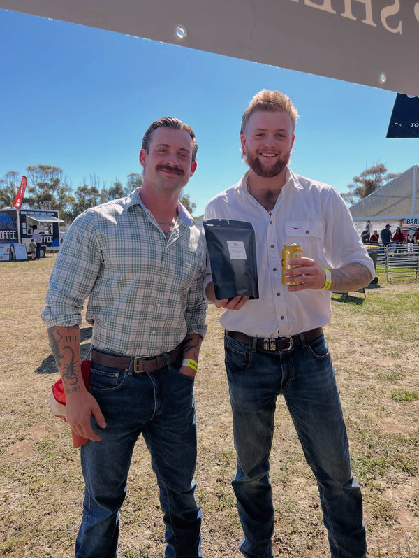 Two men standing outdoors, one holding a book and the other a can, with a clear blue sky in the background.
