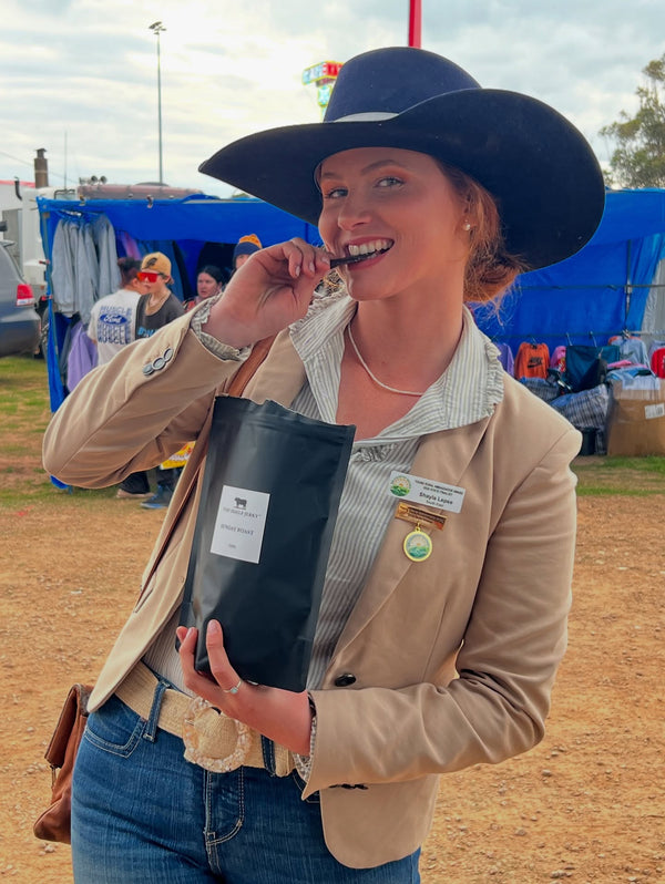 Woman in a blue hat holding a black bag with a label, standing outdoors with tents and people in the background.