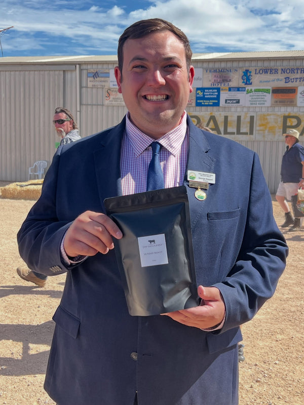 Man in a suit holding a black bag outdoors with a building and people in the background